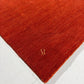 A close-up of a plush, textured area rug in a warm rust red color, showing its corner with a small gold embroidered monogram logo on a white floor.