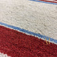 Close-up of a textured rug with a cream-colored field, a diagonal red section, and thin blue stripe accents.