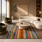 A colorful striped area rug in a modern, sunlit living room with a curved cream sofa, olive green armchairs, and a round stone coffee table. The rug features vertical stripes in blue, red, orange, yellow, green, and pink on a cream base.