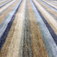 A close-up view of a textured area rug with vertical stripes in beige, brown, gray, blue, and navy, featuring a small gold monogram logo near the bottom center.