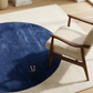 Blue circular rug with a gold logo on a white rug, accompanied by a wooden chair.