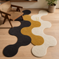 A wavy, bone-shaped area rug in three colors: black, mustard yellow, and cream. The cream section features a gold embroidered letter 'R'.