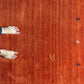 A top-down view of a warm rust-orange textured rug with visible wood grain, featuring a small gold monogram 'AR' near the center and small dark decorative motifs along the right edge. A person's bare feet are visible at the left edge for scale.