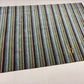 A rectangular area rug with vertical stripes in navy, green, beige, and gray, laid out on a light-colored tiled floor. A gold 'AR' logo is printed on the bottom right corner of the rug.