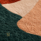 Close-up of a textured area rug with a geometric color-block pattern in dark green, peach, cream, and rust orange, featuring a gold 'AR' logo in the lower-left corner.