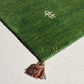 A close-up of a green textured rug corner with a multicolored tassel and small embroidered monogram details, laid on a light neutral surface.