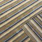 A close-up of a striped woven fabric with diagonal bands in beige, tan, brown, muted blue-gray, and off-white, showing the textile texture and a small gold embroidered logo near the lower right corner.