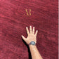 A close-up of a deep burgundy red textured rug with a gold 'AR' logo in the upper center. A person's hand and forearm, wearing a watch, are placed on the rug to show its texture and scale.
