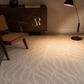 A beige area rug with a wavy, organic pattern in a styled room setting featuring a mid-century wooden chair, a dark wood credenza with a gold logo, and a floor lamp.