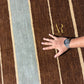 A close-up of a brown area rug with vertical stripes in beige and a muted blue-gray, shown with a person's hand and wrist resting on the surface. A gold 'R' logo is printed on the rug.