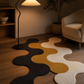 A modern wavy-pattern area rug in black, mustard yellow, and cream, styled on a wooden floor in a cozy living room setting next to a floor lamp and armchair.