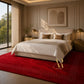 A luxury bedroom scene featuring a vibrant red area rug with a dotted geometric border, styled with a cream and beige bed, gold bedside tables, and warm ambient lighting. The brand name 'Artisia Rugs' is watermarked in gold on the bottom right corner of the rug.