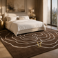 A large brown area rug with a white abstract floral or spiral pattern and a small gold logo, styled in a modern, sunlit bedroom next to a bed with white linens.