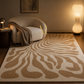 A large area rug with a beige and cream zebra-like wavy pattern, styled in a cozy living room next to a curved armchair and a glowing floor lamp.