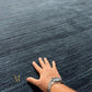 A close-up of a dark blue-gray textured rug with a human hand placed on it for scale, along with a small gold monogram logo in the lower left corner.