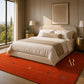 A styled bedroom scene featuring a large, plush red area rug with subtle embroidered motifs, positioned under a bed with neutral beige bedding and accessories. The room has elegant beige walls, gold bedside tables, and a large window on the left showing green trees outside.