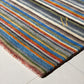 A close-up of a striped area rug with multicolored wool-like fibers, featuring diagonal bands of blue, gray, beige, orange, green, and brown, laid on a light tiled floor.