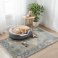 Dog resting in a gray pet bed on a decorative rug with a small table and plant in the background.