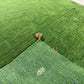 Green artificial turf or grass-like flooring material shown in a close-up, with a folded section revealing the backing and a small white monogram logo.