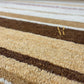 Close-up of a striped area rug with textured loops in beige, cream, and dark brown. A gold 'Arts & Lofts' logo is visible on the right side.