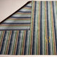 A striped area rug shown from an overhead angle, with one corner folded back to reveal the backing. The rug features vertical stripes in navy, green, beige, and brown, with a gold monogram logo in the lower right corner.