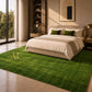 A large green textured area rug with a gold monogram logo, styled in a modern, sunlit bedroom next to a bed with neutral bedding.