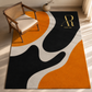 A modern area rug with an abstract wavy pattern in black, orange, and off-white, styled in a bright room next to a wooden armchair. The rug features a gold monogram logo for 'Artista Rugs' in the top right corner.