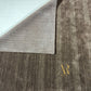 A close-up of a textured area rug in a dark brown and gray heathered pattern, with one corner folded back to show the underside. A gold 'AR' logo is printed on the lower right.