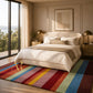 A large rectangular area rug with bold horizontal stripes in red, orange, yellow, green, blue, and purple, styled in a modern, sunlit bedroom with a cream bed and wooden nightstands.