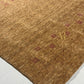 A close-up of a textured area rug in a warm camel brown color, featuring a subtle woven pattern with small burgundy diamond motifs and a gold monogram letter 'R' near the center. The rug is shown laid out on a light-colored floor.