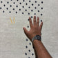 A close-up of a cream-colored textured rug with a black geometric pattern, featuring a person's hand and arm resting on it for scale, with a gold monogram watermark in the upper left.