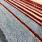 A close-up of a textured area rug with a pattern of light blue, cream, and dark red stripes, featuring a gold 'AR' logo in the top right corner.