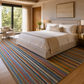 A large rectangular area rug with multicolored diagonal stripes in blue, red, orange, yellow, green, and beige, styled in a modern, sunlit hotel bedroom next to a bed with neutral bedding.