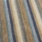 A close-up of a striped woven fabric with diagonal bands in beige, tan, brown, muted blue-gray, and off-white, showing the textile texture and a small gold embroidered logo near the lower right corner.