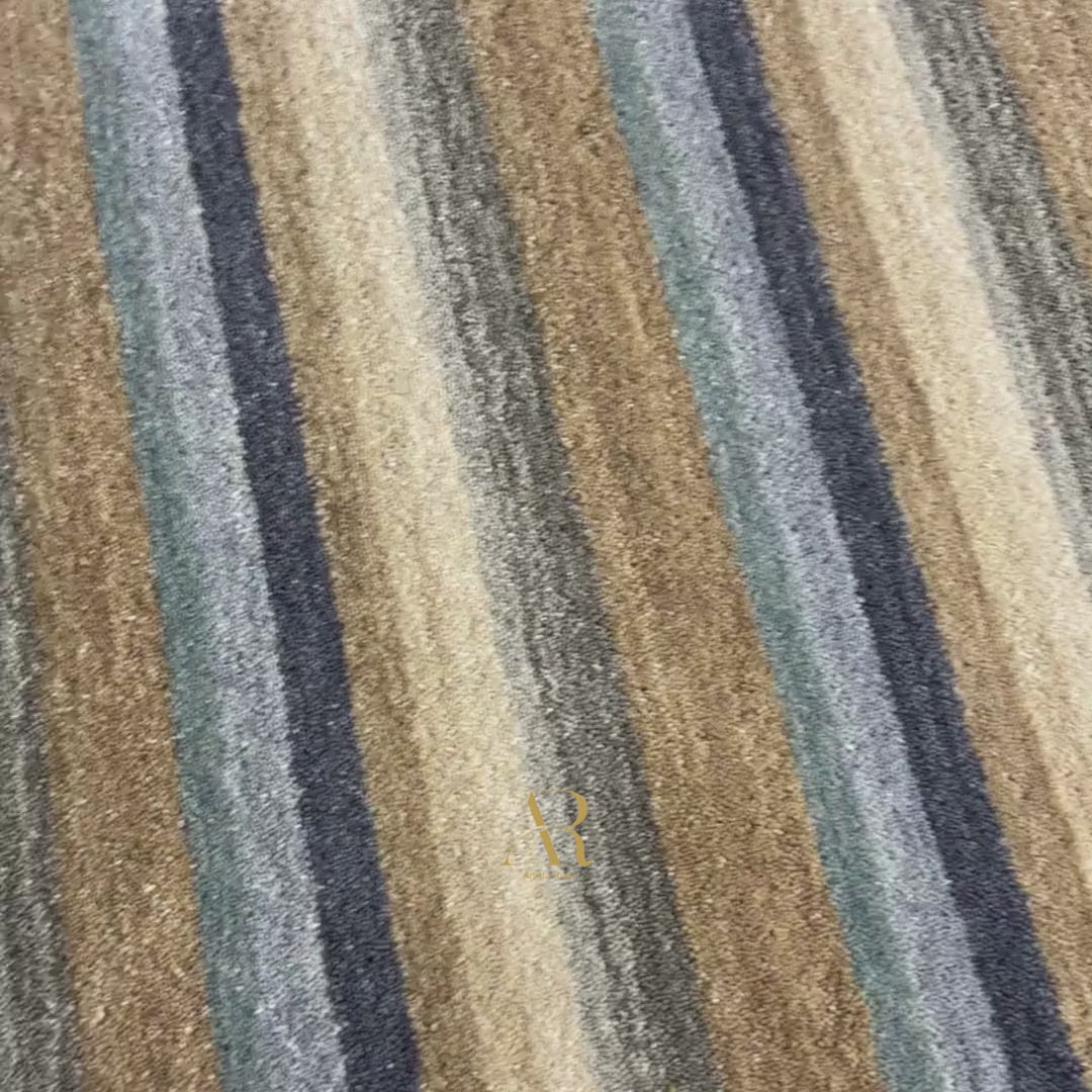 A close-up of a striped woven fabric with diagonal bands in beige, tan, brown, muted blue-gray, and off-white, showing the textile texture and a small gold embroidered logo near the lower right corner.