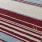 A rectangular area rug with horizontal stripes in shades of blue, cream, and burgundy, featuring a gold monogram logo in the top-left corner.