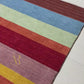 A rectangular area rug with vertical stripes in multiple colors including brown, blue, red, orange, pink, yellow, green, and light blue, featuring a gold embroidered monogram logo in the bottom left corner.