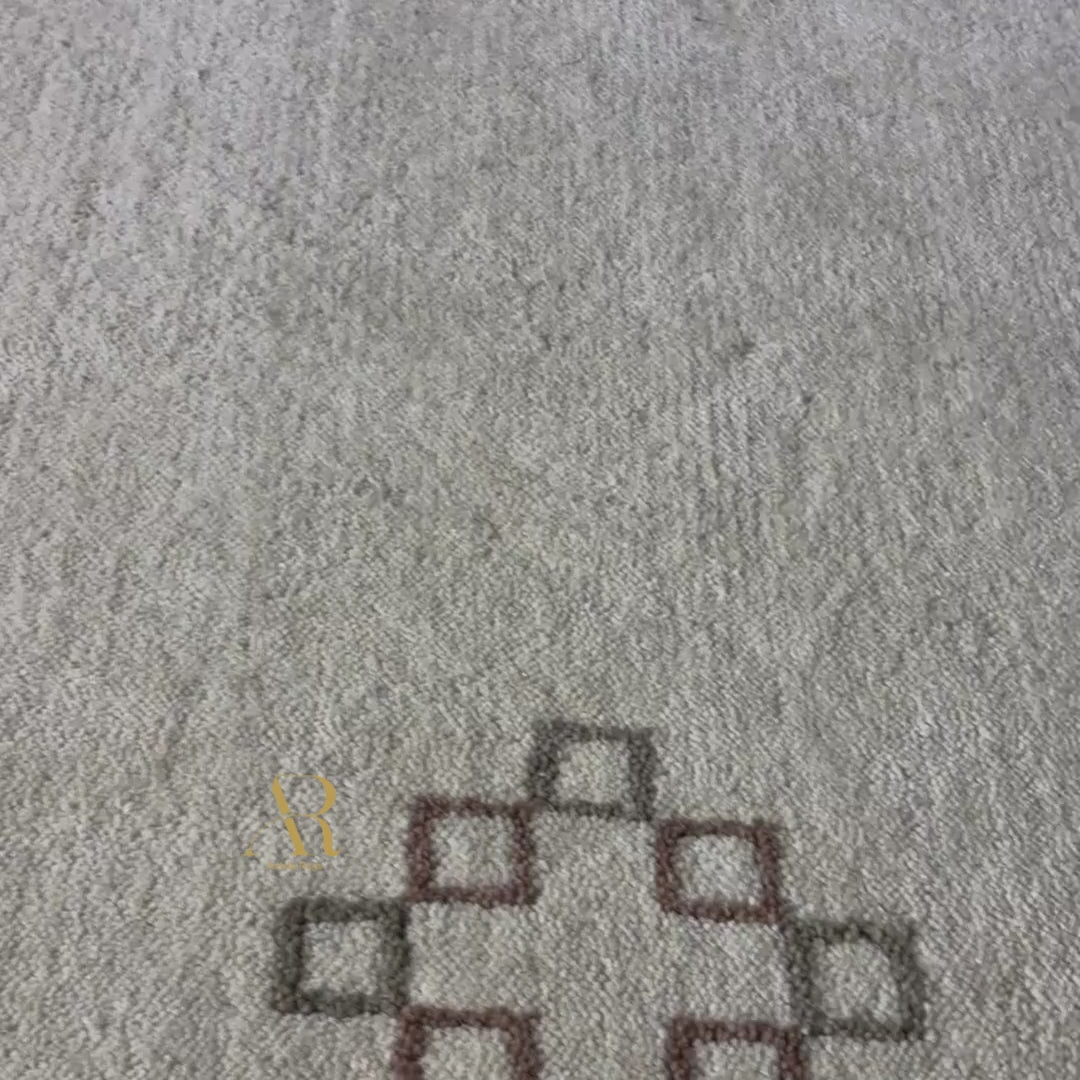 A rectangular beige area rug with a Southwestern geometric pattern in brown, gold, and black, featuring a gold monogram logo near the bottom right. The rug is shown flat against a solid white background.