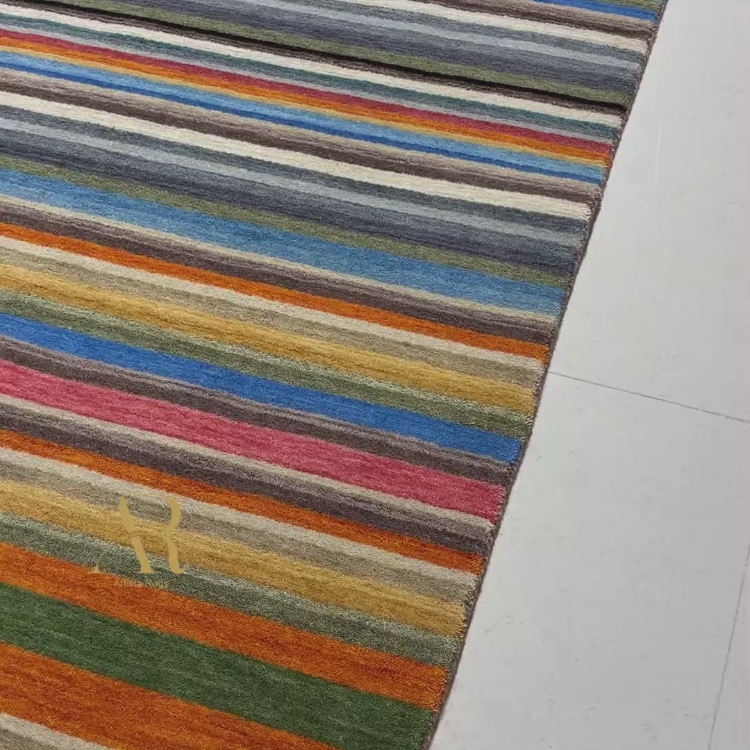 A rectangular area rug with a vibrant multicolor horizontal stripe pattern, featuring a small gold embroidered monogram logo in the upper left corner, set against a plain white background.