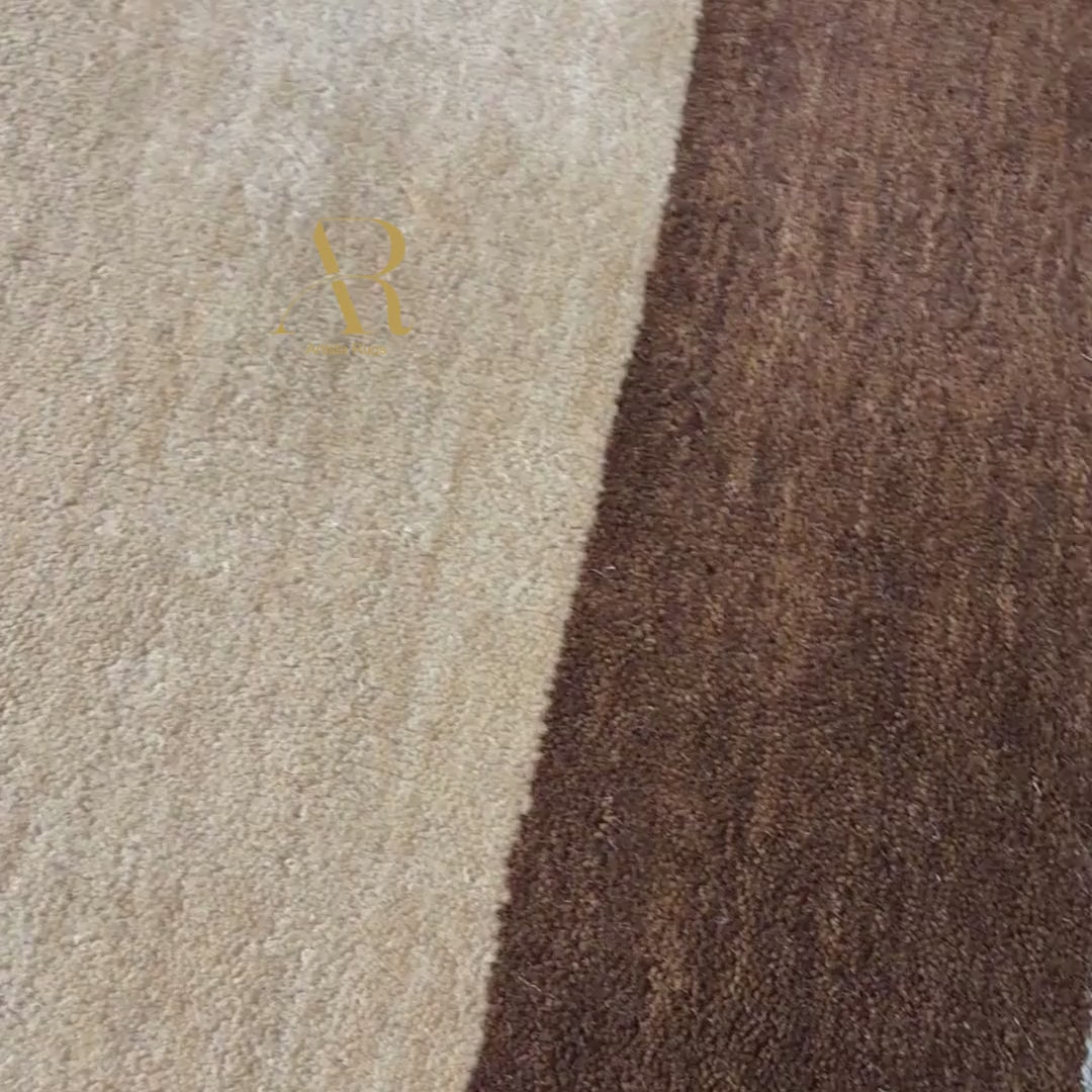 A close-up of a brown area rug with vertical stripes in beige and a muted blue-gray, shown with a person's hand and wrist resting on the surface. A gold 'R' logo is printed on the rug.