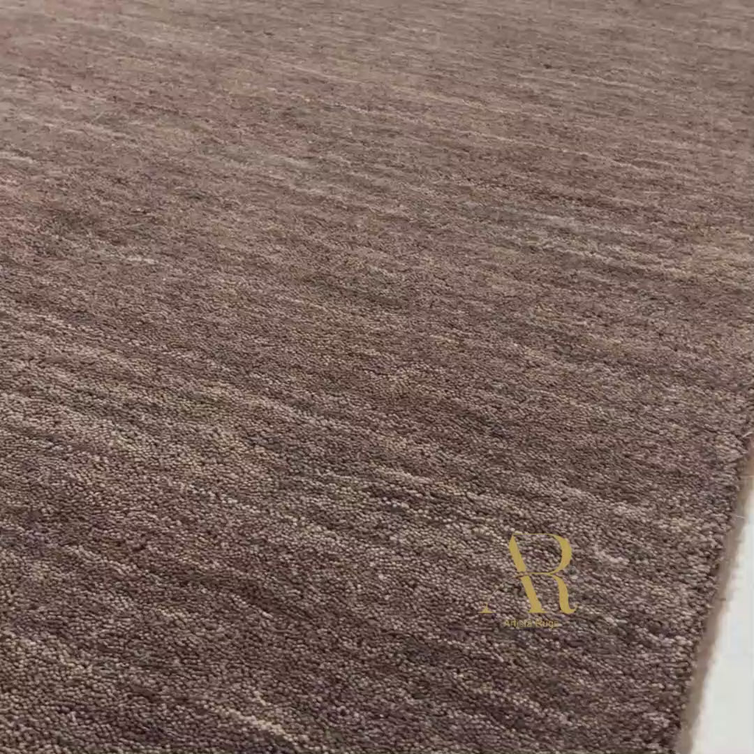 A close-up of a textured area rug in a dark brown and gray heathered pattern, with one corner folded back to show the underside. A gold 'AR' logo is printed on the lower right.