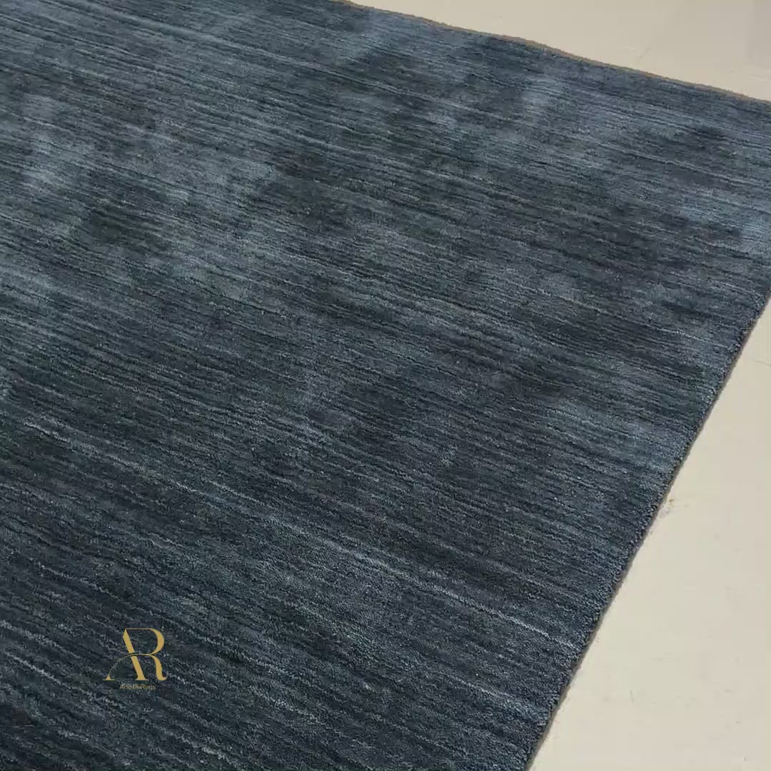 A dark navy blue area rug with a subtle horizontal textured pattern and a small gold monogram logo in the bottom right corner, shown against a plain white background.