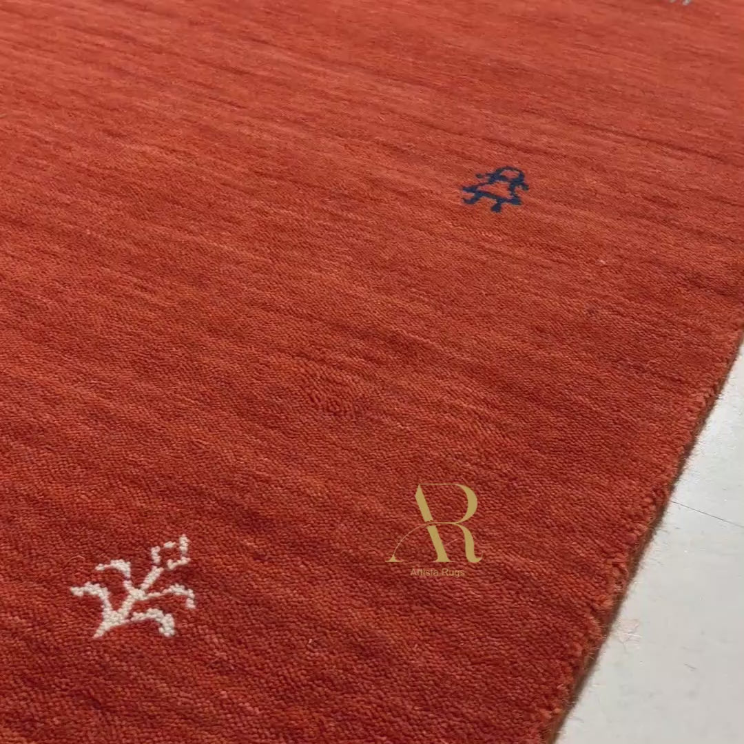 A rectangular red woven rug with a subtle vertical texture, featuring a gold 'AR' logo in the center, accented with small black and white geometric details around the border, shown on a plain white background.