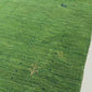 Green artificial turf or grass-like flooring material shown in a close-up, with a folded section revealing the backing and a small white monogram logo.