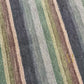 A rectangular area rug with vertical stripes in multiple colors including brown, blue, red, orange, pink, yellow, green, and light blue, featuring a gold embroidered monogram logo in the bottom left corner.