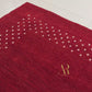 A red rectangular rug with a decorative border of small white dots and a gold monogram in the upper right corner, shown flat against a white background.