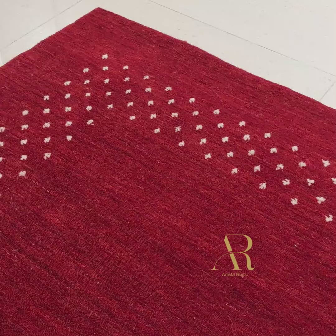 A red rectangular rug with a decorative border of small white dots and a gold monogram in the upper right corner, shown flat against a white background.