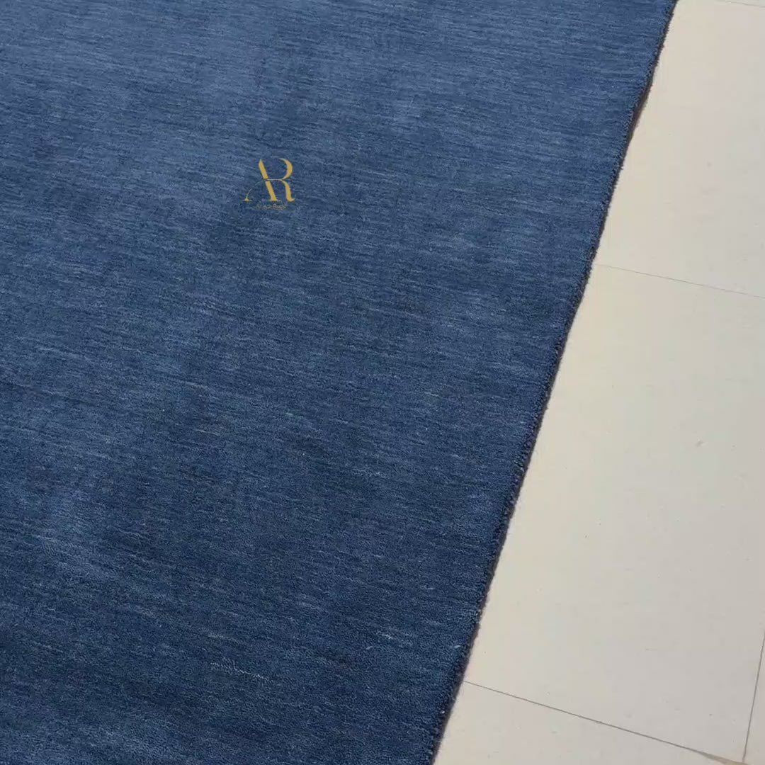 A rectangular royal blue textured rug with a small gold embroidered monogram logo in the upper right quadrant, shown flat against a plain white background.