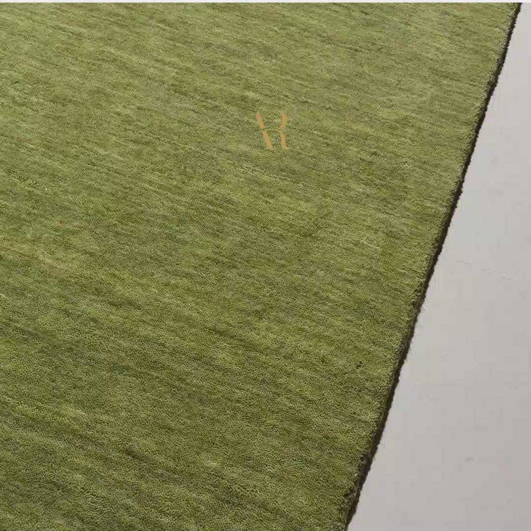 An olive green textured area rug with a small gold monogram logo in the lower right corner, displayed flat against a white background.