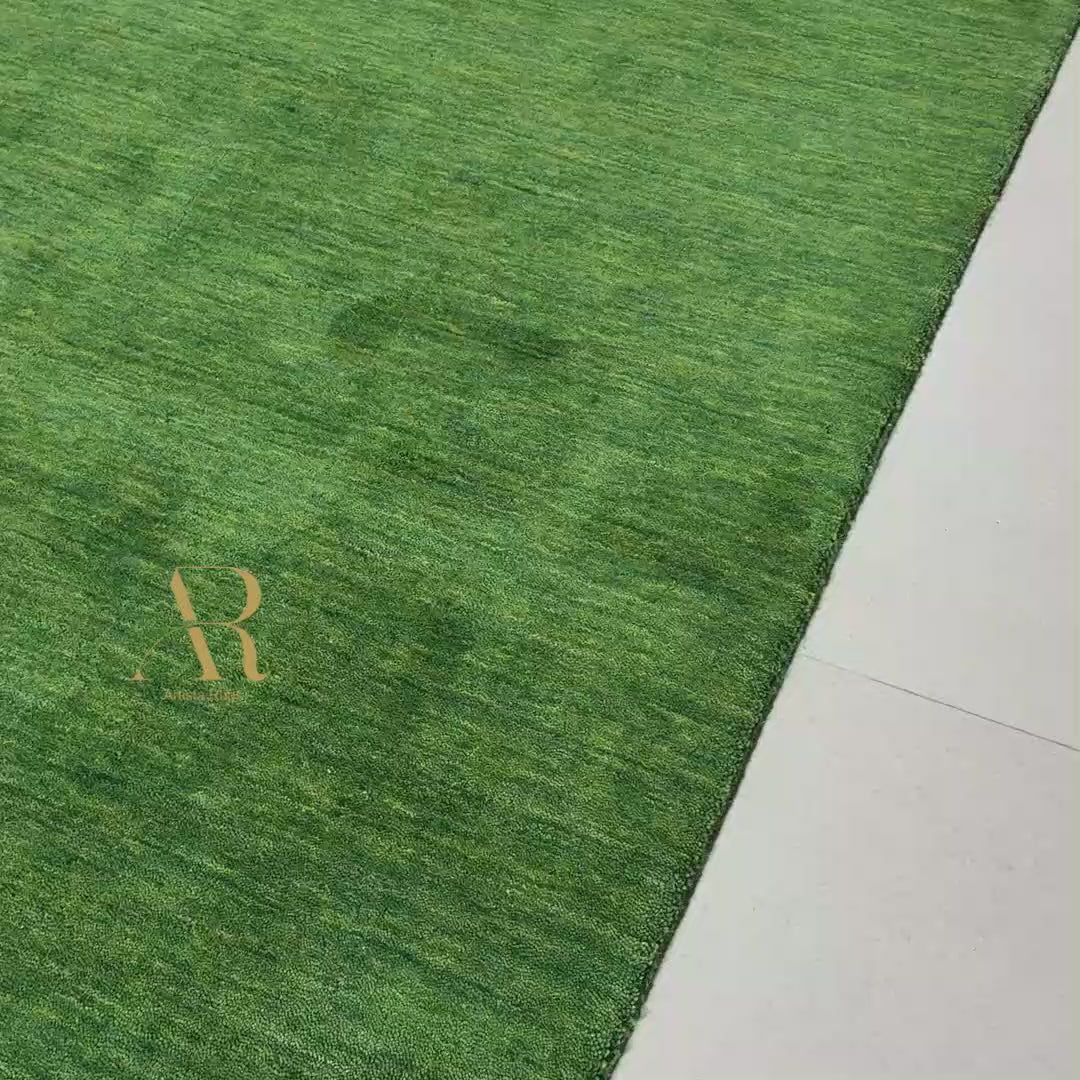 A rectangular green artificial turf or grass mat with a textured grass-like surface, laid out on a light-colored tiled floor. A gold monogram logo reading 'AR' is printed near one corner.